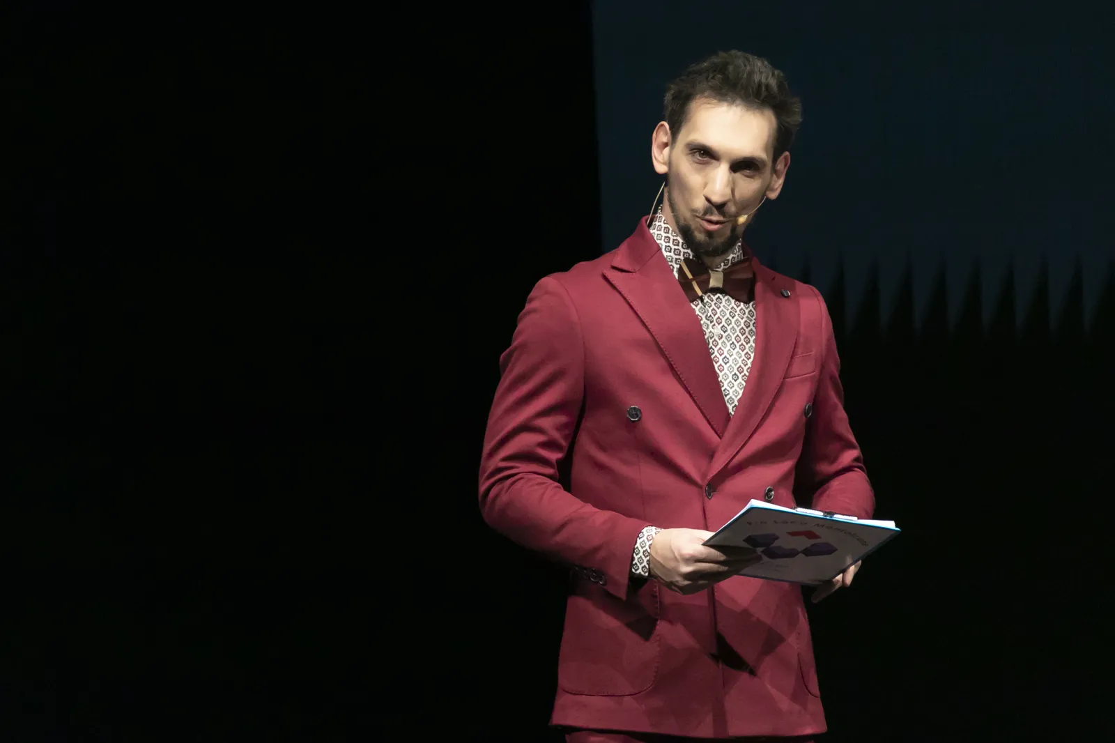 Ritratto di Nicola Valletta sul palco con libretto in mano, microfono ad archetto, abito bordeaux